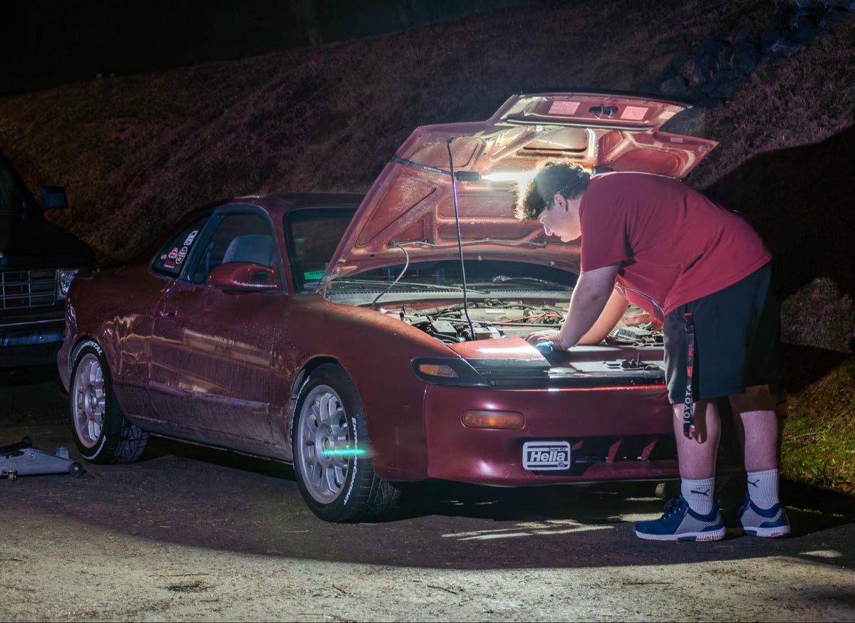 Person working on a red car with the hood open at night. Engine bay illuminated by a FLEXIT Underhood light.