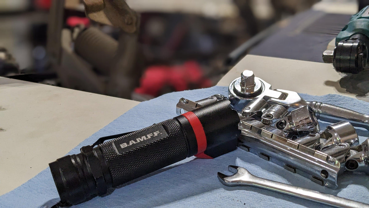 BAMFF 8.0 Tactical Flashlight sitting on a blue shop towel with some sockets, socket wrench, and a box end wrench. Blurred out car engine bay in the background.